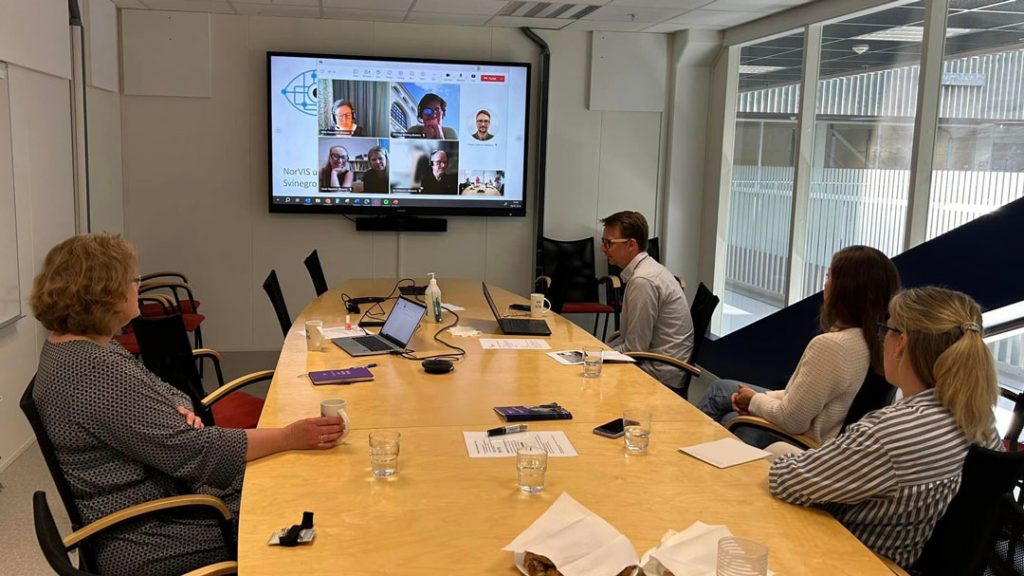 Fire av konferansedeltakerne sitter rundt et bord og snakker med deltakere som deltar digitalt på skjerm.